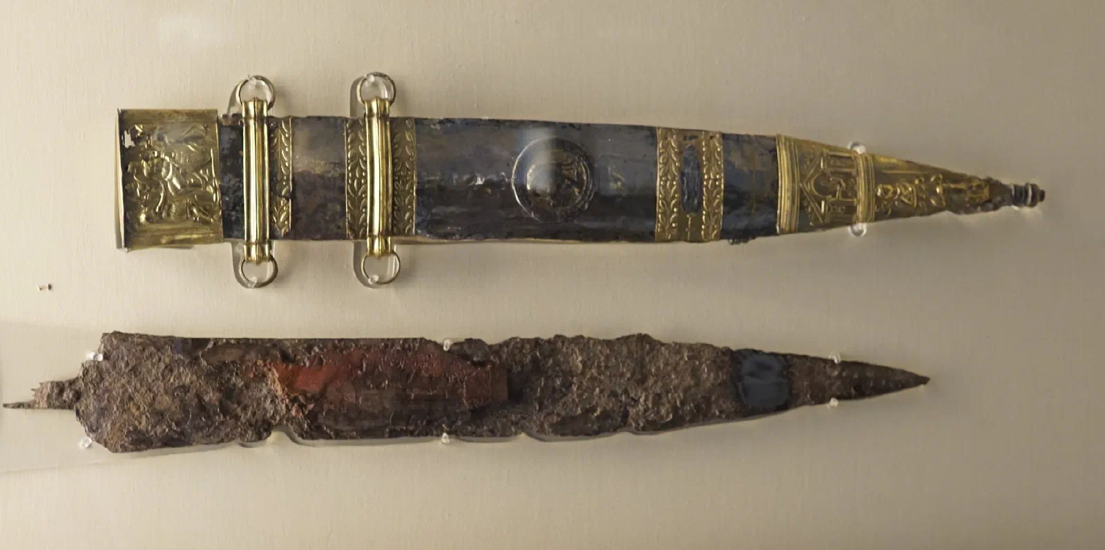 A corroded iron sword blade below its ornate gilt-bronze scabbard, decorated with a raised relief scene.