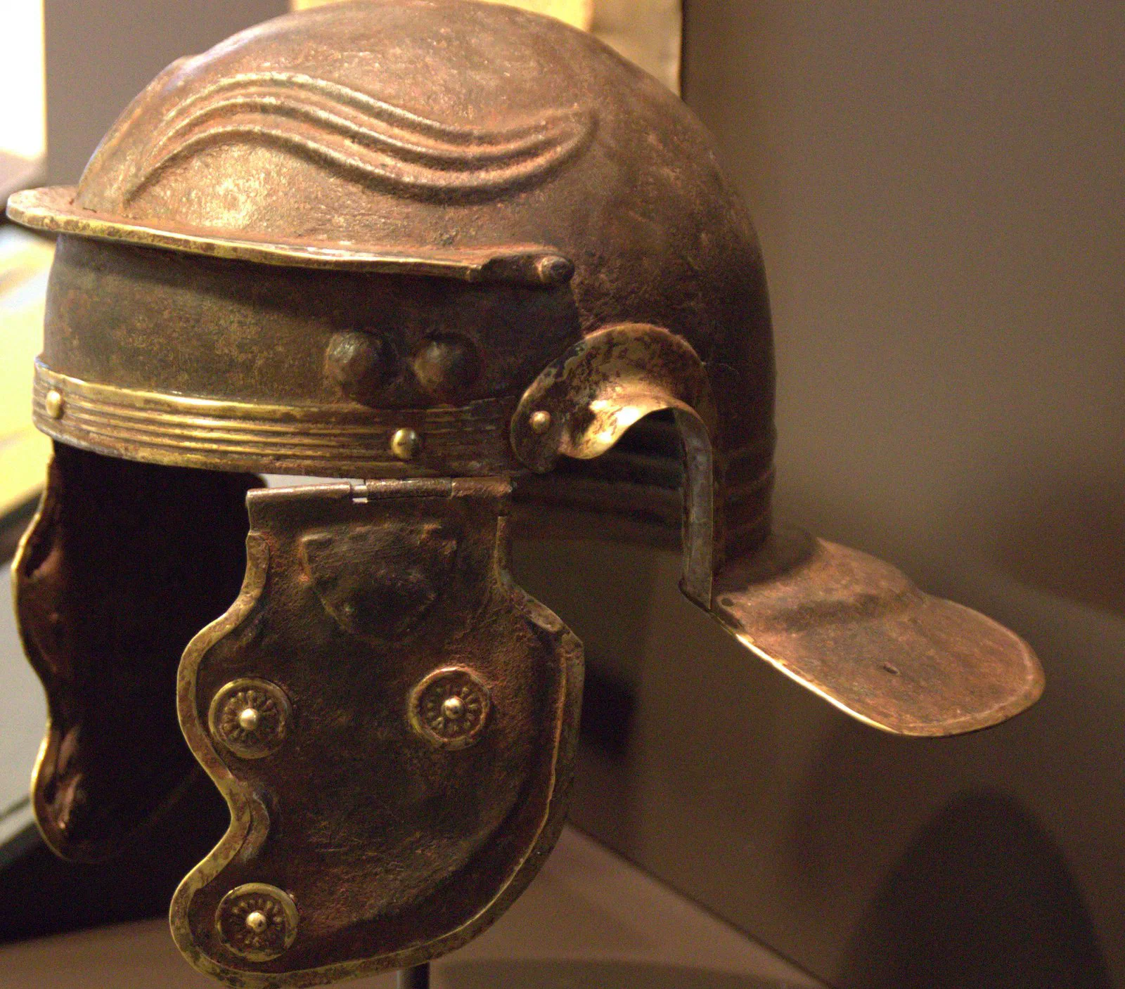 A corroded iron Roman helmet with embossed brow ridges, brass trim, one hinged cheek piece, and a wide flared neck guard.