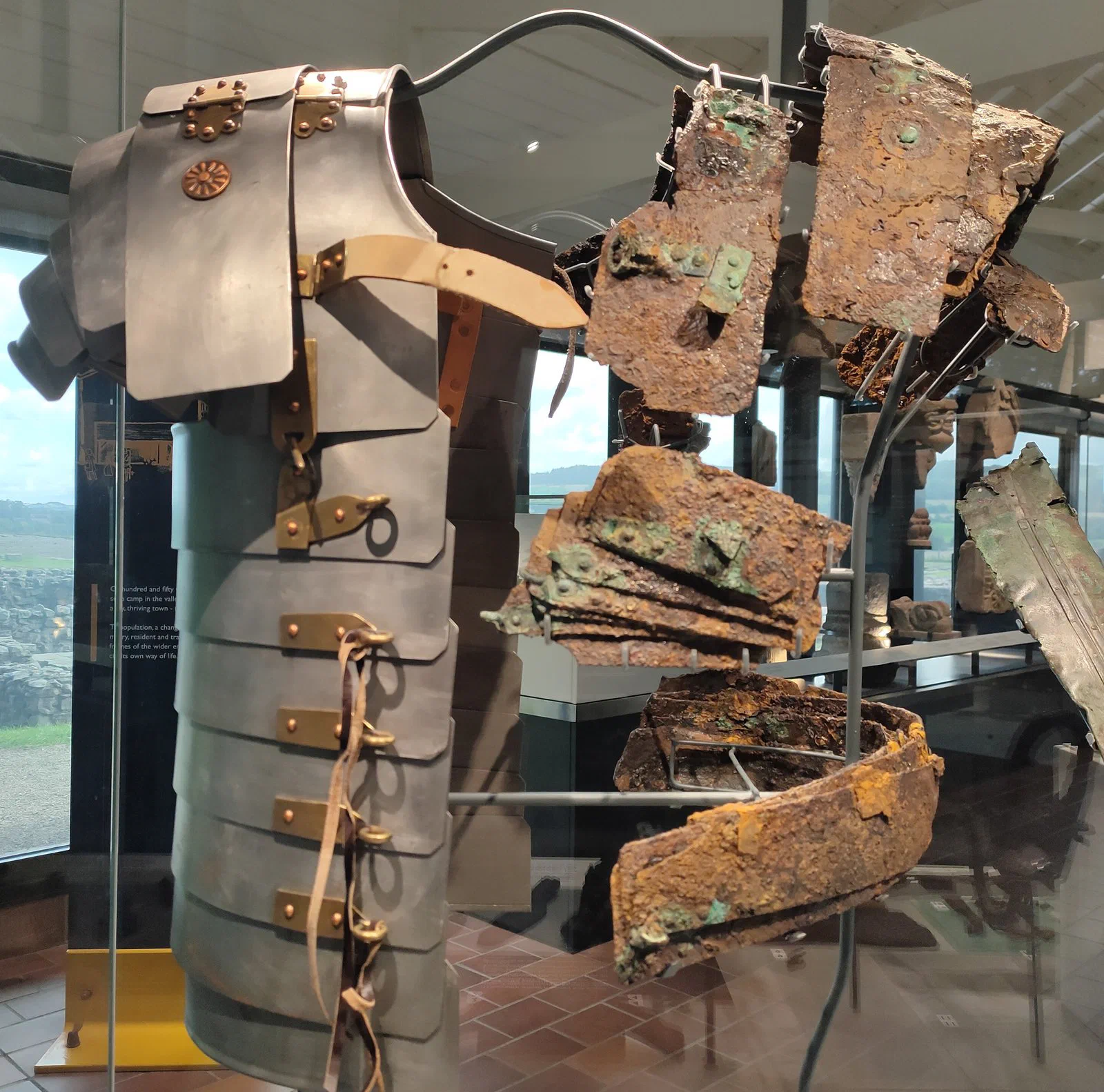 A museum case: a shiny modern reconstruction of banded plate armour beside the corroded iron fragments it was based on.